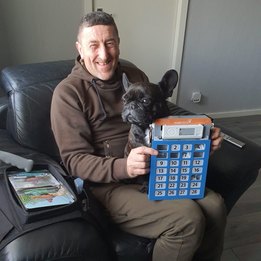 Happy YOURmeds user in an armchair with his dog, holding up the YOURmeds medication pack